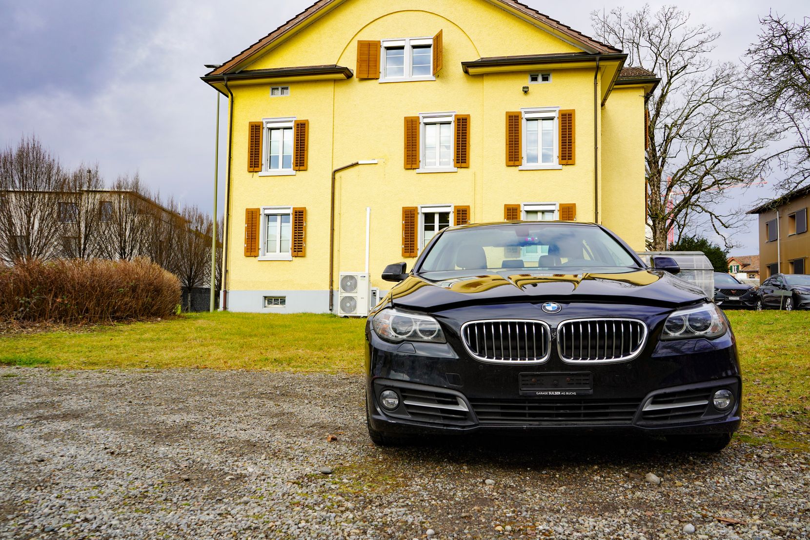 Man sieht einen schwarzen BMW auf einem Kiesplatz vor einem zweistöckigen Haus.